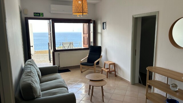 Living room with 3 seater sofa and armchair, leading to small balcony and sea view.